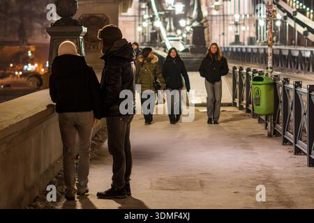 Fußgänger, die nachts im Winter entlang der Széchenyi-Kettenbrücke spazieren, werden in einer offenen Straßenszene festgehalten, die das tägliche städtische Leben zeigt. Stockfoto