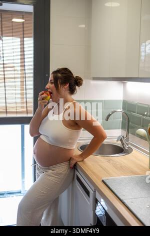 In einer stilvollen, lichtdurchfluteten Küche nimmt eine schwangere Frau einen Biss von einem saftigen Apfel und genießt dabei einen gesunden Snack, während sie sich entspannt. Ihr Glück strahlt in dieser gemütlichen Umgebung aus. Stockfoto