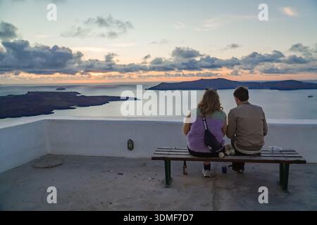 Junges Paar, das auf einer Bank sitzt und den Sonnenuntergang über der vulkanischen Caldera in Fira (Thira), Santorin, Griechenland, beobachtet. Romantische Reiseszene. Stockfoto