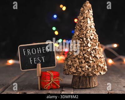 Kleine Tafel mit der deutschen Aufschrift Frohe Weihnachten auf einem Holztisch, verschwommene Weihnachtslichter im Hintergrund Stockfoto