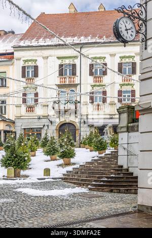 Eine festliche Winterszene auf einem europäischen Stadtplatz mit Schnee, Weihnachtsbäumen im Topf, Lichterketten, einer kunstvollen Fassade und einer großen Uhr auf einem Dekor Stockfoto