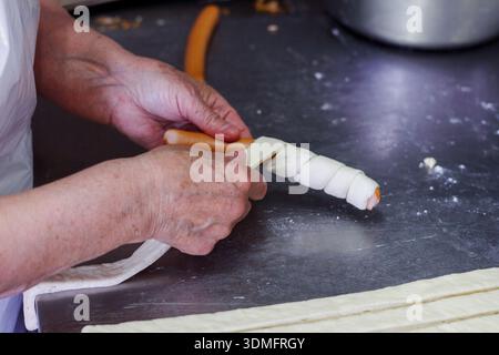 Nahaufnahme eines professionellen Küchenchefs, der Teig um eine Wurst wälzt, um frisches Gebäck in einer kommerziellen Bäckerei zuzubereiten. Stockfoto