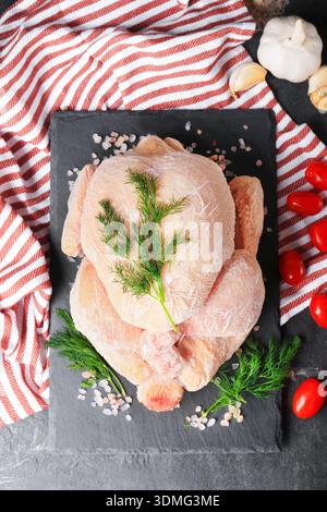 Blick von oben auf das ganze gefrorene Huhn auf schwarzem Schieferbrett mit Dill und Tomaten. Rohes Geflügelfleisch mit Frost, bereit zum Kochen auf dunklem Tisch. Hoch Stockfoto