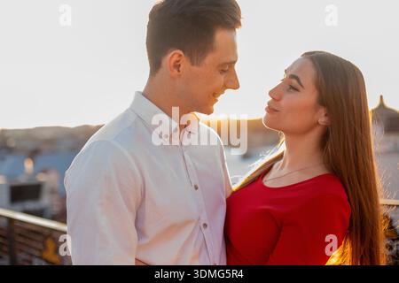 Ein Paar steht nahe auf einem Dach während des Sonnenuntergangs in der Stadt. Sie teilen ein Lächeln und schauen sich gegenseitig in die Augen, wenn die Sonne untergeht. Stockfoto