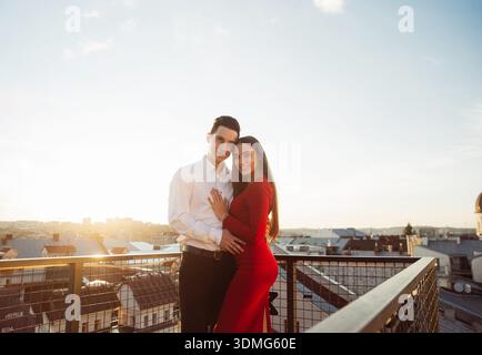 Ein Paar genießt einen Moment auf einem Dach mit Blick auf die Stadt, während die Sonne hinter sich untergeht und eine warme Atmosphäre schafft. Stockfoto