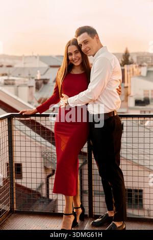Zwei Personen stehen nahe auf einem Balkon mit Blick auf die Dächer der Stadt bei Sonnenuntergang. Sie lächeln und genießen den gemeinsamen Moment. Stockfoto