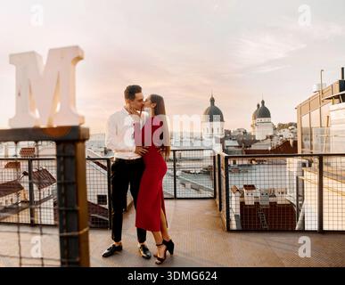 Ein Paar teilt sich einen Kuss auf einem Dach in Madrid, während die Sonne über der Stadt untergeht, mit Blick auf die Skyline und dem Buchstaben M in der Nähe. Stockfoto