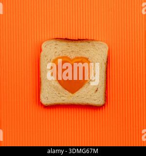 Ei aus herzförmigem Eigelb und geröstetem Brot auf orangefarbenem Hintergrund. Flache Lagen. Love-Konzept. Stockfoto