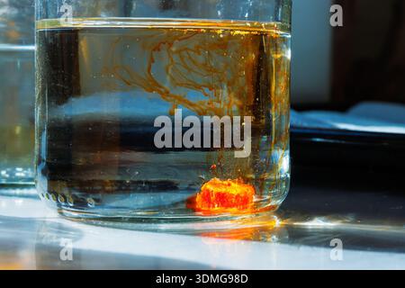 Sonnendurchflutete Nahaufnahme eines Glasbehälters mit Wasser und orangefarbenem Lebensmittelfarbstoff, der sich im Wasserbehälter löst und farbenfrohe Diffusionslinien im Sonnenlicht erzeugt Stockfoto