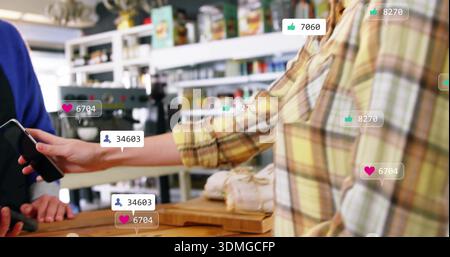 Kunde in gelbem Karomuster, der sein Telefon am Café-Theke präsentiert, mit umwickelten Broten und sozialen Symbolen Stockfoto