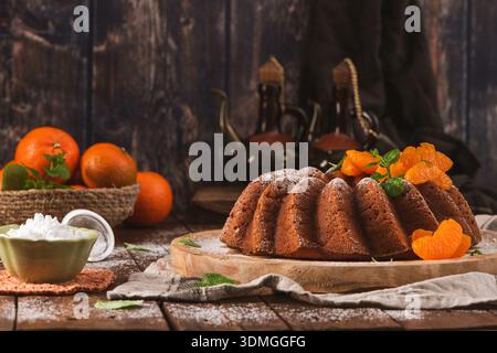 Ein goldbrauner Zitruskuchen mit frischen Mandarinensegmenten und Minze steht auf einem rustikalen Holztisch. Die Szene bietet ein warmes, stimmungsvolles Licht Stockfoto