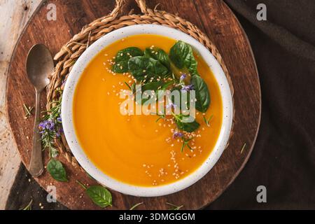 In einer weißen Schüssel wird ein lebhaftes orangenes Gemüsepüree serviert, garniert mit frischen Spinatblättern und Sesamsamen. Die rustikale Kopfaufnahme zeigt wa Stockfoto