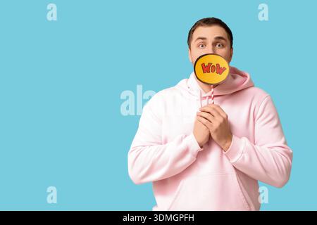 Schockierter junger Mann mit Sprechblase mit Wort WOW auf blauem Hintergrund Stockfoto