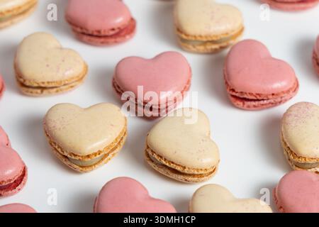 Makronen sind in einem Raster auf einer weißen Oberfläche angeordnet. Diese Leckereien haben die Form von Herzen und sind in zwei Farben erhältlich: Rosa und Creme. Stockfoto