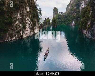 Erkunden Sie die ruhige Schönheit des Cheow Lan Lake, während ein hölzernes Boot durch smaragdgrüne Gewässer gleitet, umgeben von hohen Kalksteinklippen und üppigem Grün Stockfoto
