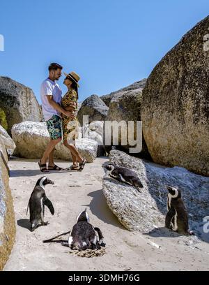 Ein Paar genießt einen romantischen Moment am Sandstrand von Boulders Beach. Afrikanische Pinguine bewegen sich in der Nähe in dieser atemberaubenden natürlichen Umgebung und machen es zu einem der besten Stockfoto