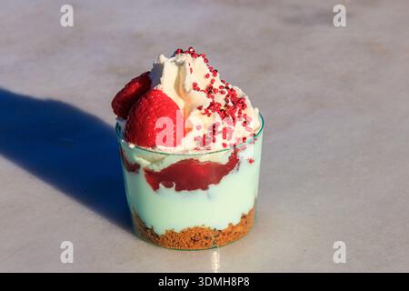 Erdbeer-Kleinigkeiten mit Schlagsahne, Beerensauce und Kekskrümeln in einem Glas auf grauem Betonhintergrund Stockfoto