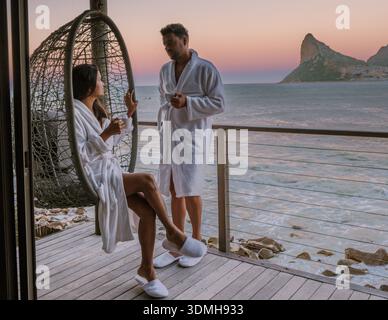 Ein Paar in gemütlichen Bademänteln genießt dampfende Tassen Kaffee, während er den atemberaubenden Sonnenaufgang über dem Meer in Kapstadt, Südafrika, bestaunt. Stockfoto