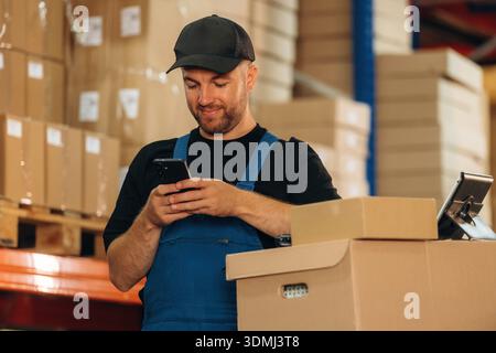 SMS per Telefon. Mann in Uniform arbeitet im Lager mit Paketen für den Versand. Stockfoto