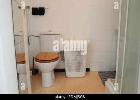 Weißes Badezimmer mit Toilette mit Holzsitzbezug und weißem Wäschekorb. Saubere Alltagsszene, die sich auf einfaches Badezimmer konzentriert Stockfoto