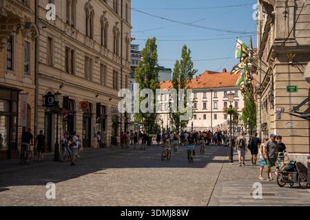 Ljubljana, Slowenien - 10. August 2025: Eine sonnenverwöhnte Kopfsteinpflasterstraße in Ljubljana voller Menschen, die an historischen europäischen Bu vorbeilaufen und Rad fahren Stockfoto