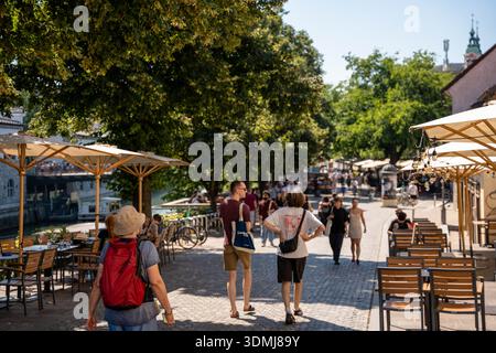 Ljubljana, Slowenien - 10. August 2025: Sonnendurchflutete Pflasterpromenade in Ljubljana an einem Fluss mit Café-Sitzgelegenheiten unter großem Sonnenschirm Stockfoto