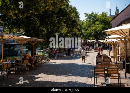 Ljubljana, Slowenien - 10. August 2025: Lebhafter Sommertag in Ljubljana entlang des Flusses Ljubljanica, wo Touristen einen Kopfsteinpflasterweg entlang schlendern Stockfoto