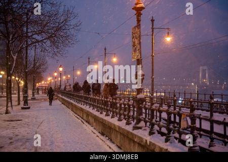 Budapest, Ungarn - 6. Januar 2026: In der Abenddämmerung spazieren die Menschen entlang einer verschneiten Uferpromenade in Budapest, wo leuchtende Vintage-Straßenlaternen und ein wr Stockfoto