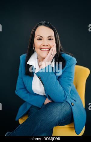 Lächelnde Profi-Frau in einem Blazer, die auf einem gelben Stuhl sitzt Stockfoto