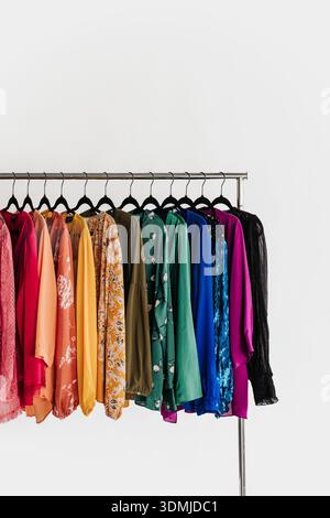 Die Oberteile für Damen hängen in Regenbogenreihenfolge vor weißem Hintergrund Stockfoto
