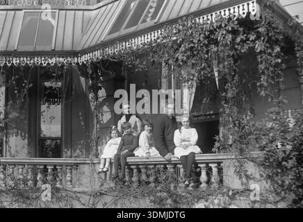 Viktorianisches Foto einer Familiengruppe, die auf dem prunkvollen Balkon einer Residenz im viktorianischen Stil posierte und wahrscheinlich aus dem späten 19. Oder frühen 20. Jahrhundert stammt. Zwei Erwachsene und vier Kinder sitzen an einer Steingeländer, eingerahmt von Kletterweben und komplexen architektonischen Details wie dekorativen Griffarbeiten und großen Glasfenstern. Stockfoto