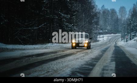 Ein Auto fährt auf einer verschneiten Winterstraße im Norden Russlands. Die Scheinwerfer leuchten durch den fallenden Schnee, mit dunklem Wald, vereisten Spuren und kaltblauem Licht Stockfoto