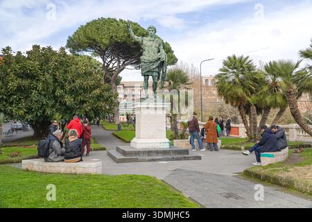 Statue einer römischen Figur, möglicherweise Julius Cäsar, in einem üppigen öffentlichen Park in Neapel, Kampanien, Italien. Menschen, die sich in der Nähe vor dem grünen Meer entspannen Stockfoto