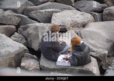 Pärchen sitzen auf großen Steinbrüchen in der Nähe von Punto panoramico Belvedere Argentina, Neapel (Neapel), Italien. Sie teilen sich Pizza zum Mitnehmen und lesen draußen Stockfoto