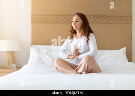 kaukasische Frau mittleren Alters, die zu Hause auf dem Bett meditiert und Achtsamkeit übt Stockfoto