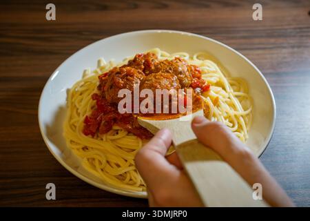 Nahaufnahme der Hände, die Tomatensauce mit Fleischbällchen mit einem Holzspachtel auf Spaghetti servieren. Stockfoto