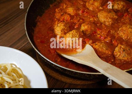 Nahaufnahme von Fleischbällchen, die in reichhaltiger Tomatensauce in einer Pfanne mit einem Holzlöffel kochen. Stockfoto
