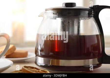 Teekanne voller schwarzem Tee auf einem Holztisch mit getrockneten Zitronenscheiben, einer Tasse und Gebäck in einem hellen Raum. Vorderansicht. Stockfoto