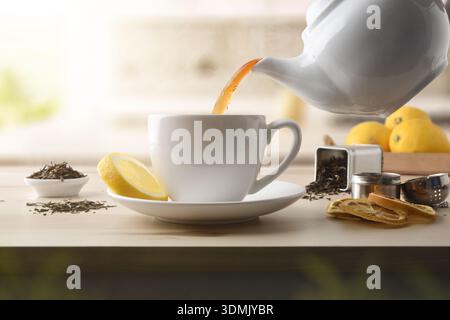 Details zum Servieren von Tee aus einer Keramik-Teekanne in eine weiße Keramikschale mit einer Zitronenscheibe auf einem Holztisch mit Zutaten und getrockneten Teeblättern. F Stockfoto