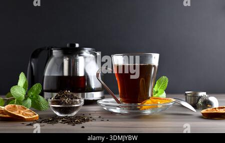 Glasbecher und Teekanne voll Tee auf einer Holzbank mit einer Schüssel getrockneter Teeblätter mit dunklem isoliertem Hintergrund. Vorderansicht. Stockfoto