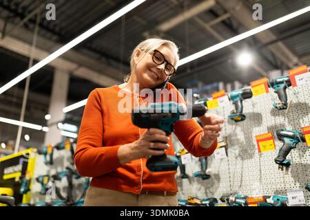 Eine lächelnde ältere Frau mit Brille steht in einem Eisenwarengeschäft, hält eine Bohrmaschine und spricht mit ihrem Telefon, während sie die Werkzeugauswahl durchsucht. Stockfoto