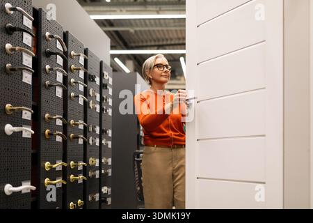 Eine Frau in einem stylischen orangefarbenen Pullover untersucht Türbeschläge in einem Heimwerkergeschäft. Sie inspiziert sorgfältig eine weiße Tür, während sie darüber nachdenkt, zu unterscheiden Stockfoto