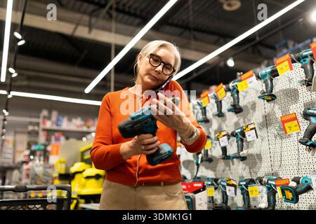 Eine Frau mit Brille ist in einem Eisenwarenladen, hält eine Übung und spricht mit ihrem Telefon. Sie prüft Optionen zwischen verschiedenen Werkzeugen im Regal und Stockfoto