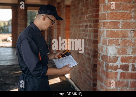 Dokumente und Abgleichwerkzeug sind in der Hand. Der Baumeister arbeitet auf der Baustelle, unvollendetes Gebäude. Stockfoto