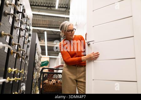 Eine Frau mit Brille und einem orangefarbenen Pullover untersucht eine weiße Tür in einem Geschäft für Heimwerker. Sie konzentriert sich auf die Merkmale der Tür in der Hardware Stockfoto