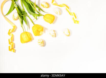 Der Blumenstrauß aus gelben Tulpen mit grünen Blättern ist neben dekorierten Ostereiern und gelbem Band auf weißem Hintergrund angeordnet und sorgt für einen festlichen Frühling Stockfoto