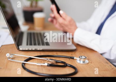 Medizinischer Fachmann überprüft das Telefon während der Arbeit am Laptop mit Stethoskop auf dem Schreibtisch in einem geschäftigen Klinikbüro Stockfoto