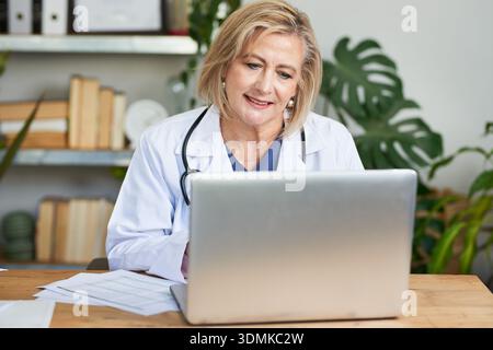 Leitende Ärztin mit Laptop am Schreibtisch mit Stethoskop und medizinischen Dokumenten im modernen Büro Stockfoto