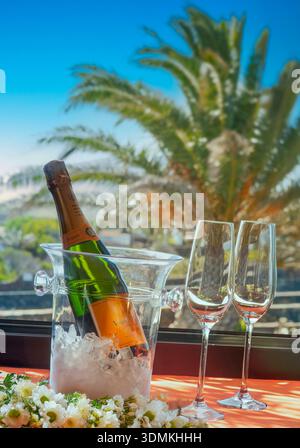 Sektflasche Blumen und Gläser auf Eis im Weinkühler auf der offenen Terrasse der luxuriösen Ferienvilla/Hotelzimmer, Sonnenpalme und blauer Himmel dahinter Stockfoto