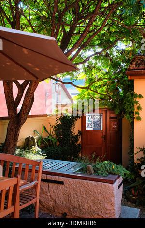 Eine ruhige und sonnige Außenterrasse mit rustikalem Holzrestaurant unter einem großen Gartenschirm. Die leuchtend rote Säule, üppig grüner Tre Stockfoto